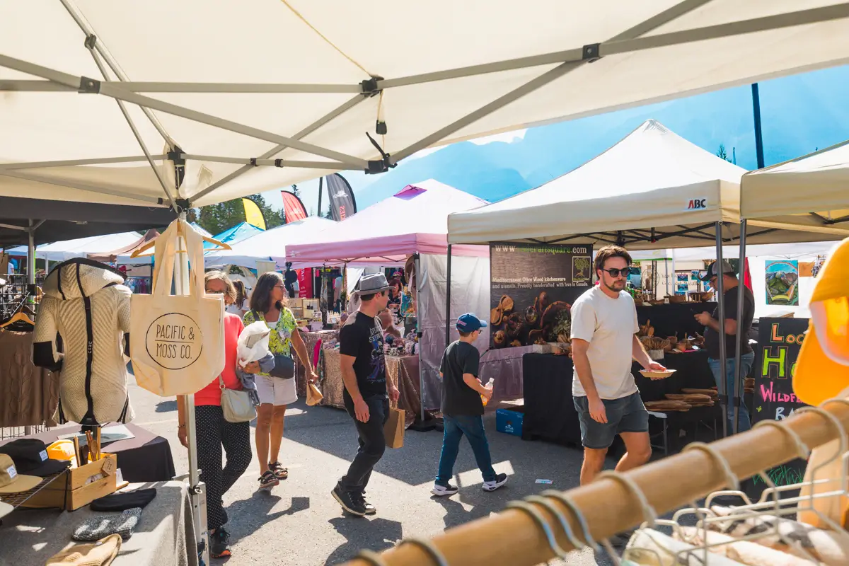 People walk among vendor tents at an outdoor market in Canmore on a sunny day. Stalls display various goods, and shoppers browse while others explore the lively market area.