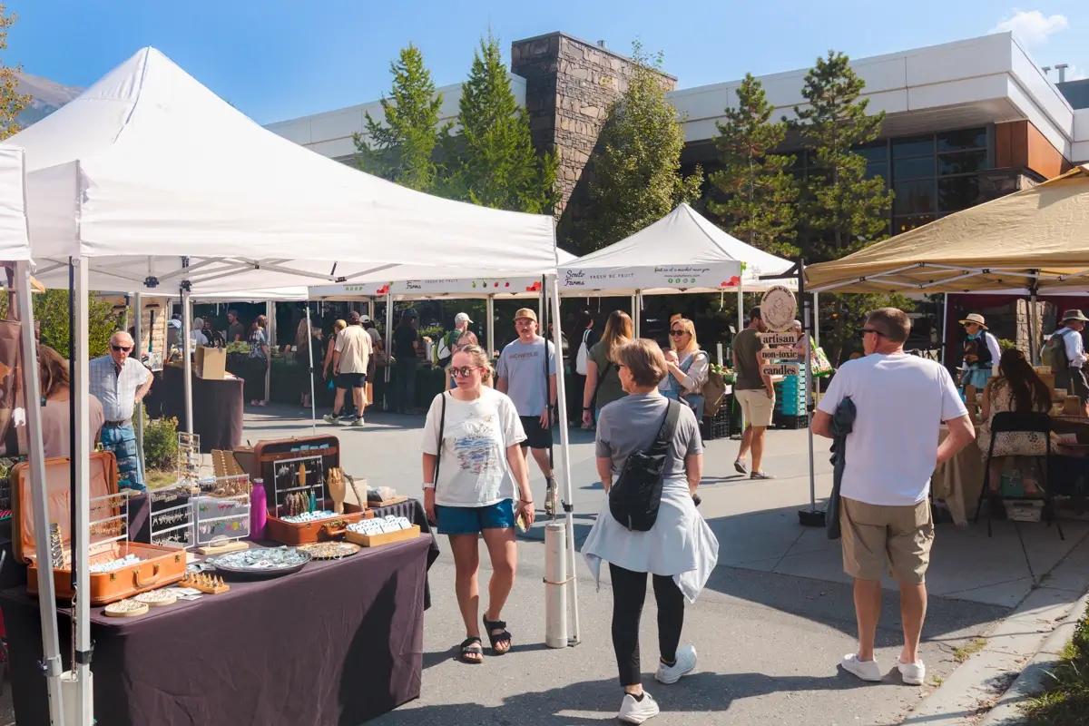 People browse vendor booths selling handmade goods at an outdoor market in Canmore on a sunny day. White canopy tents line a paved area near a modern building, all surrounded by trees.