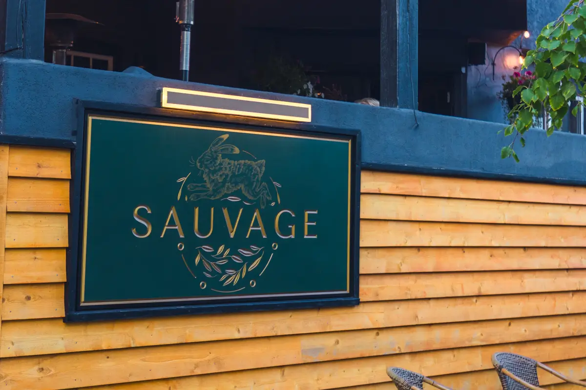 A green sign with gold text reads Sauvage and is mounted on a wall with horizontal wooden panels in Canmore. The sign features a graphic of an animal above the word and decorative elements below.