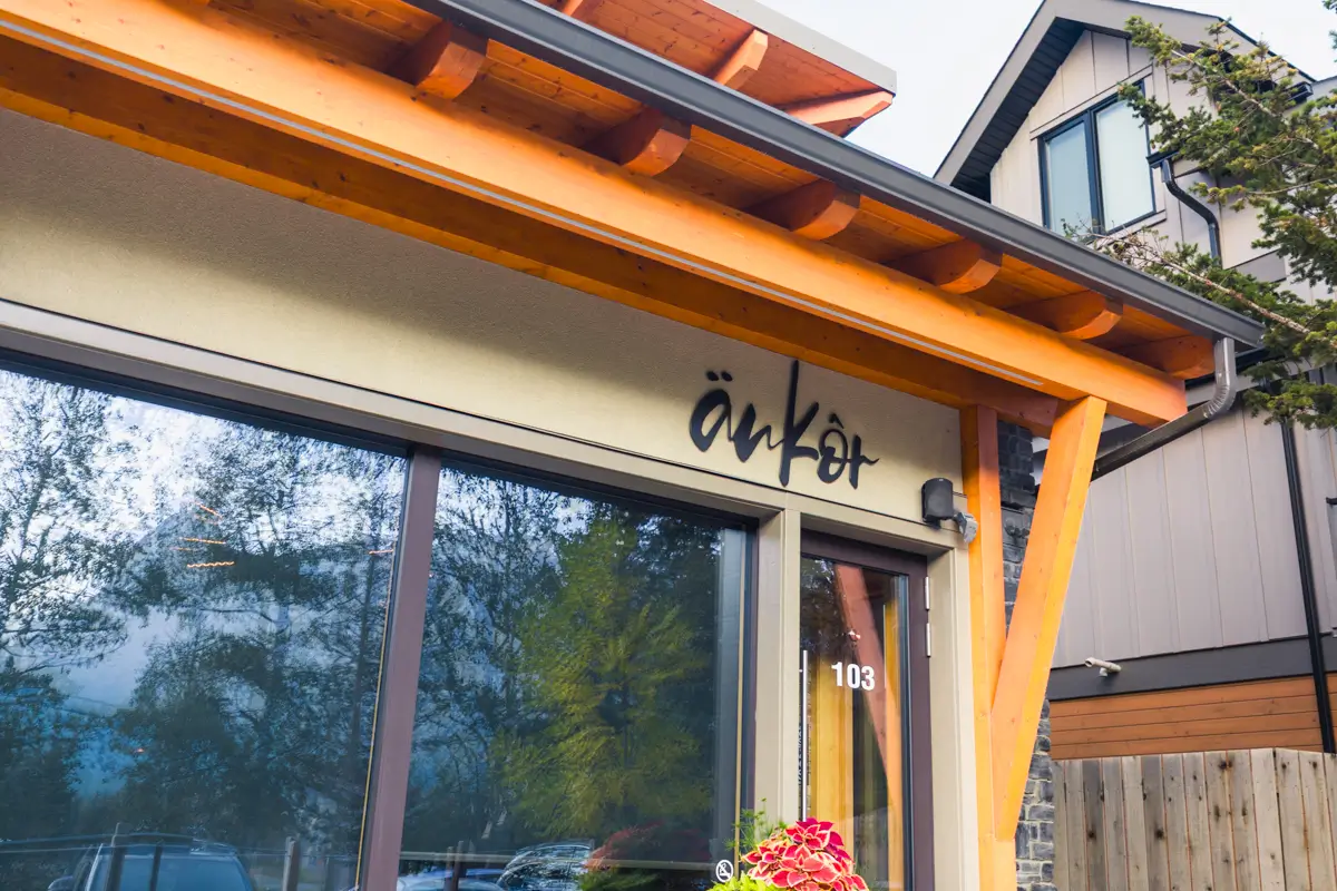 The exterior of a Canmore building with large windows and wooden beams displays the word änkôr above the entrance and the number 103 on the glass door. Trees and another house are visible in the background.
