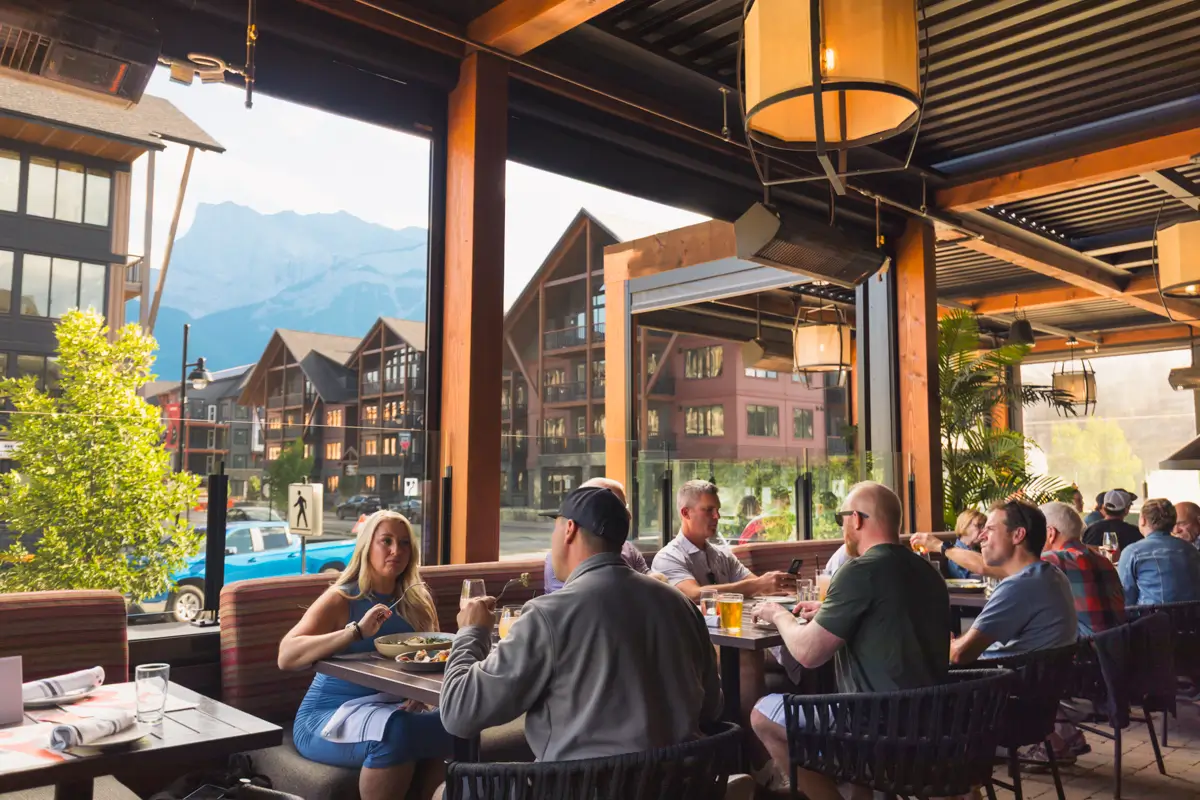 A group of people sit at tables eating and talking in a Canmore restaurant with large windows, mountain views, and chalet-style buildings visible outside.