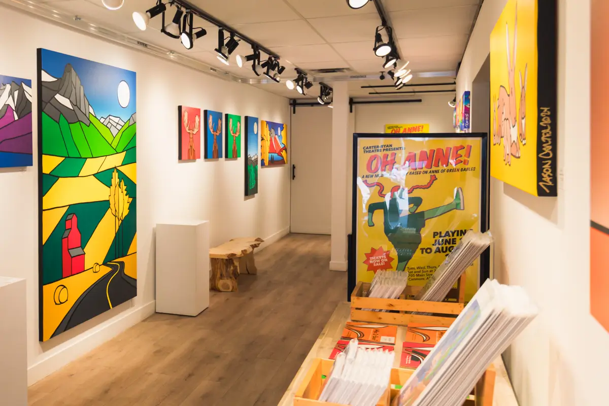 A brightly lit Canmore art gallery features colorful, modern paintings on white walls, a wooden bench, and bins filled with posters. A vibrant “Oh Anne!” poster stands out near the entrance.