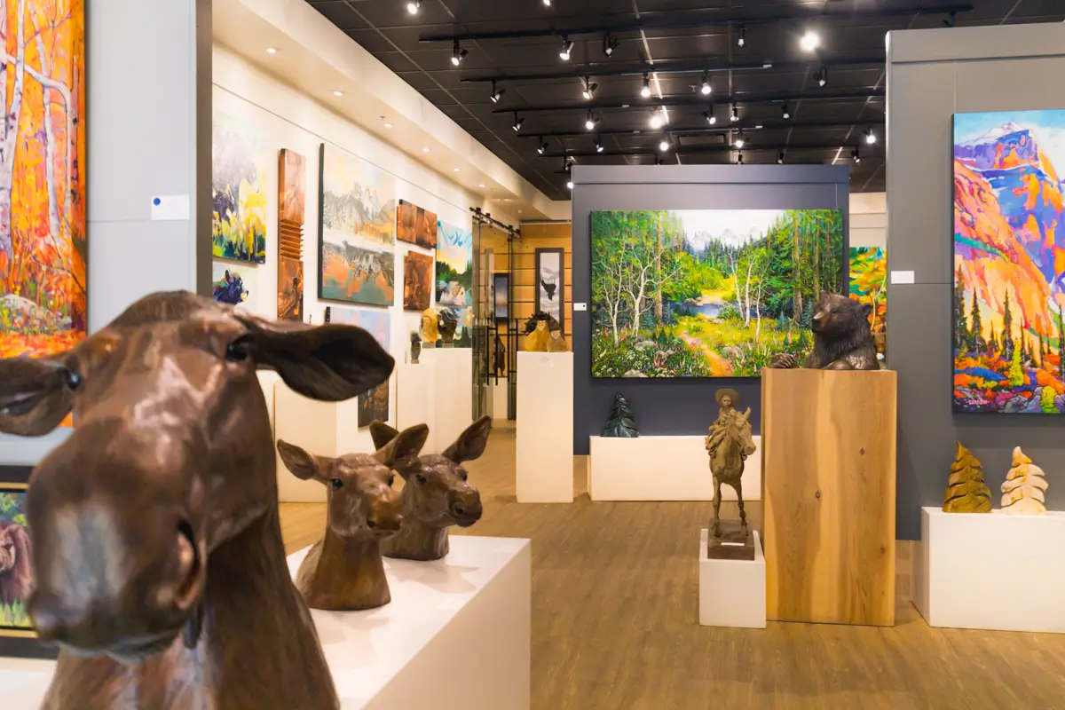 Art gallery interior in Canmore featuring animal sculptures, such as moose and deer heads in the foreground, with colorful landscape paintings of natural scenery displayed on the walls and partitions throughout the space.