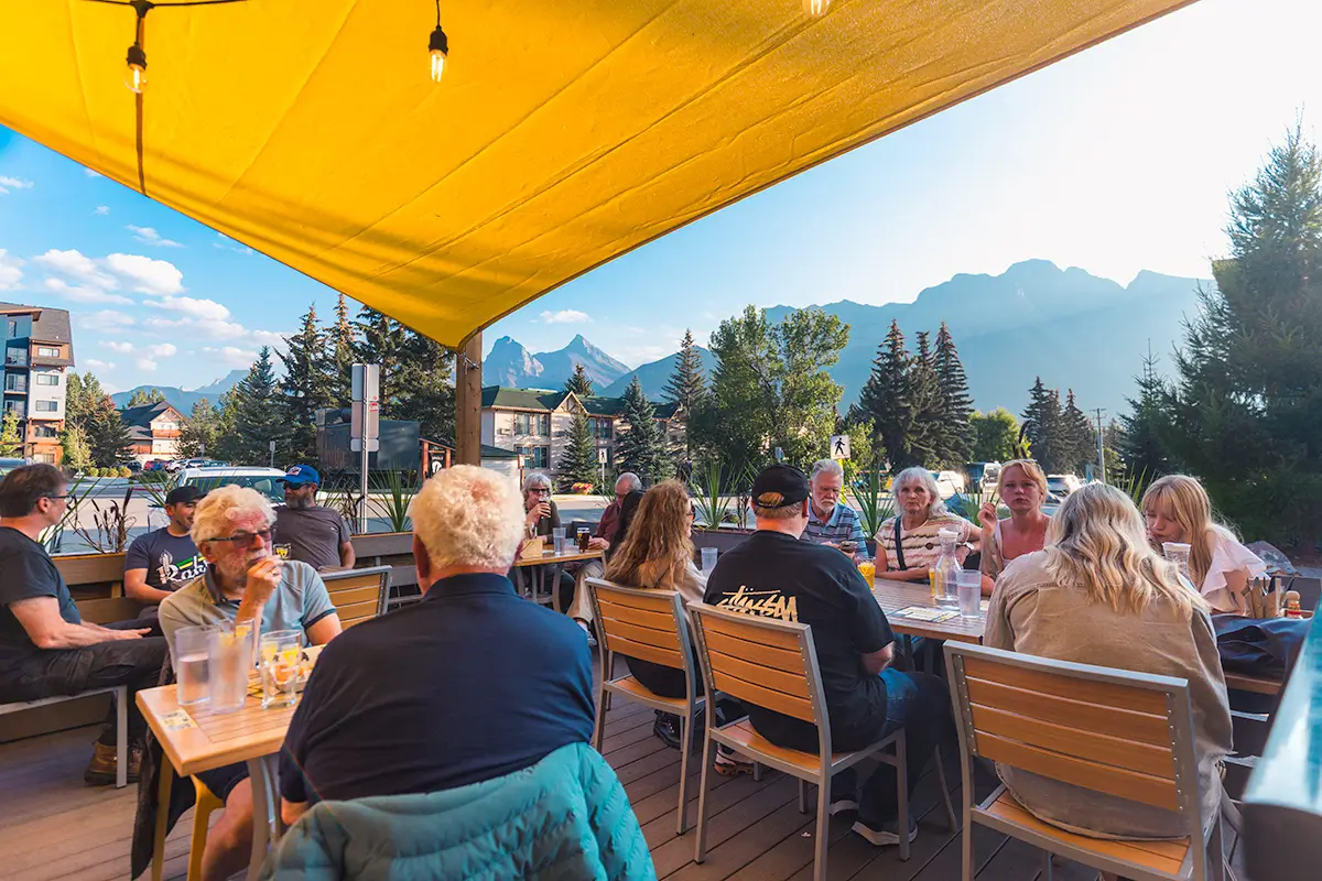 People are seated at outdoor tables under a yellow canopy, talking and dining in Canmore. The patio overlooks trees and distant mountains on a sunny day, with glasses and plates on the wooden tables.