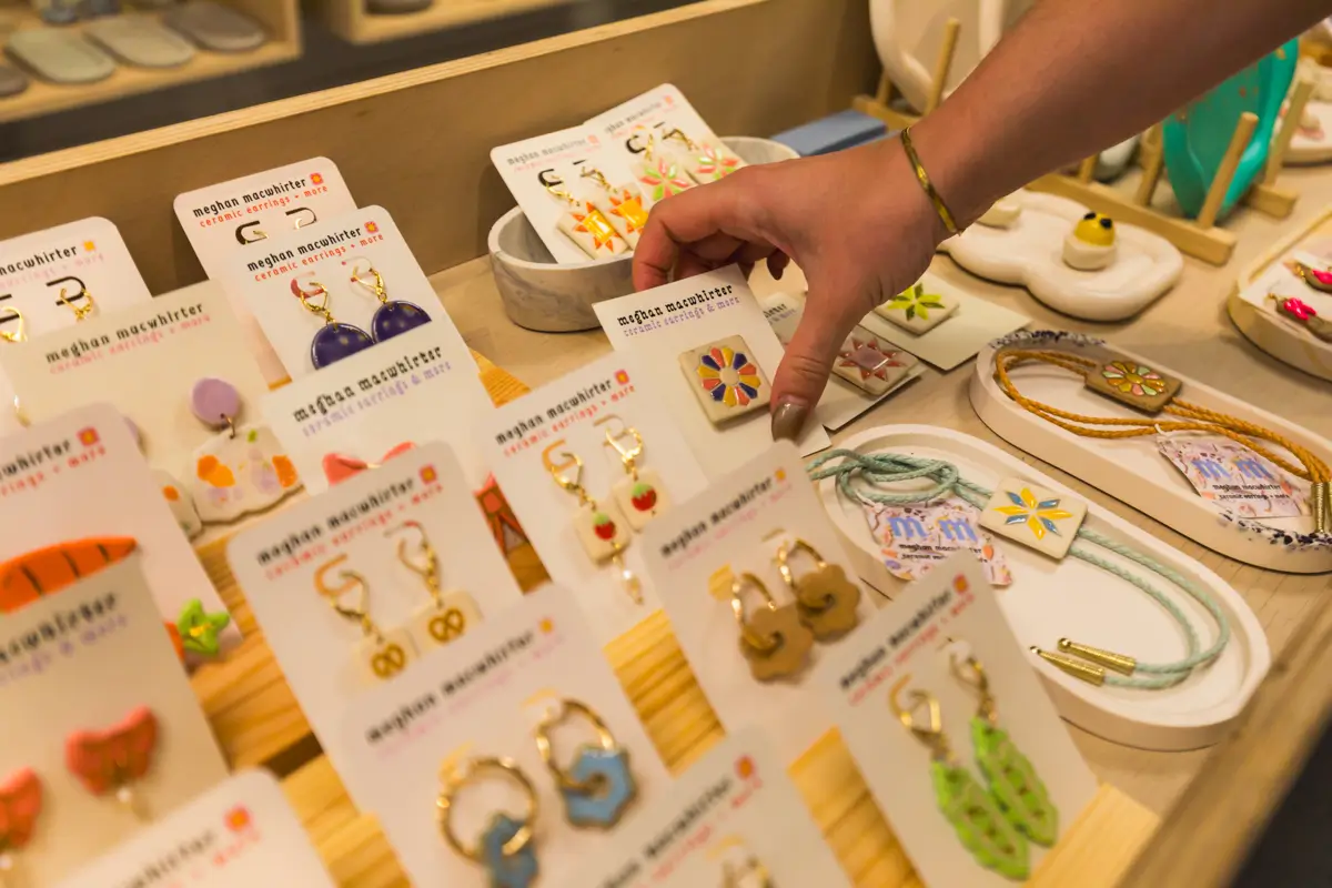 A hand reaches for a pair of colorful earrings on a display of assorted handmade earrings and jewelry, all arranged on labeled cards in a charming Canmore shop setting.