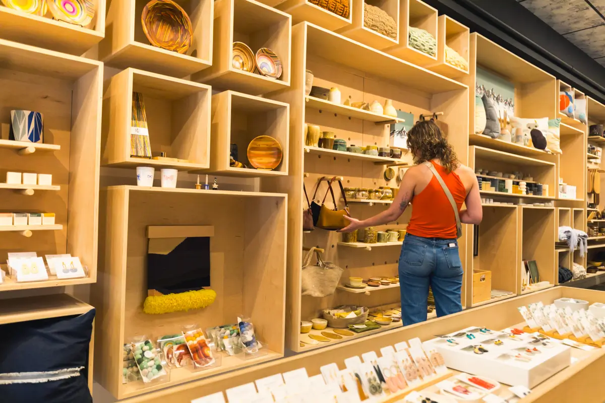 A person in a sleeveless orange top and jeans browses shelves filled with ceramics, baskets, and decorative items in a modern Canmore shop. A display counter with jewelry and small goods is in the foreground.