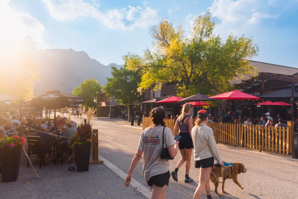 People stroll along a sunlit street in Canmore lined with outdoor restaurants and umbrellas. Two people walk a dog, others relax at tables, while trees with green and yellow leaves frame mountain views in the background.