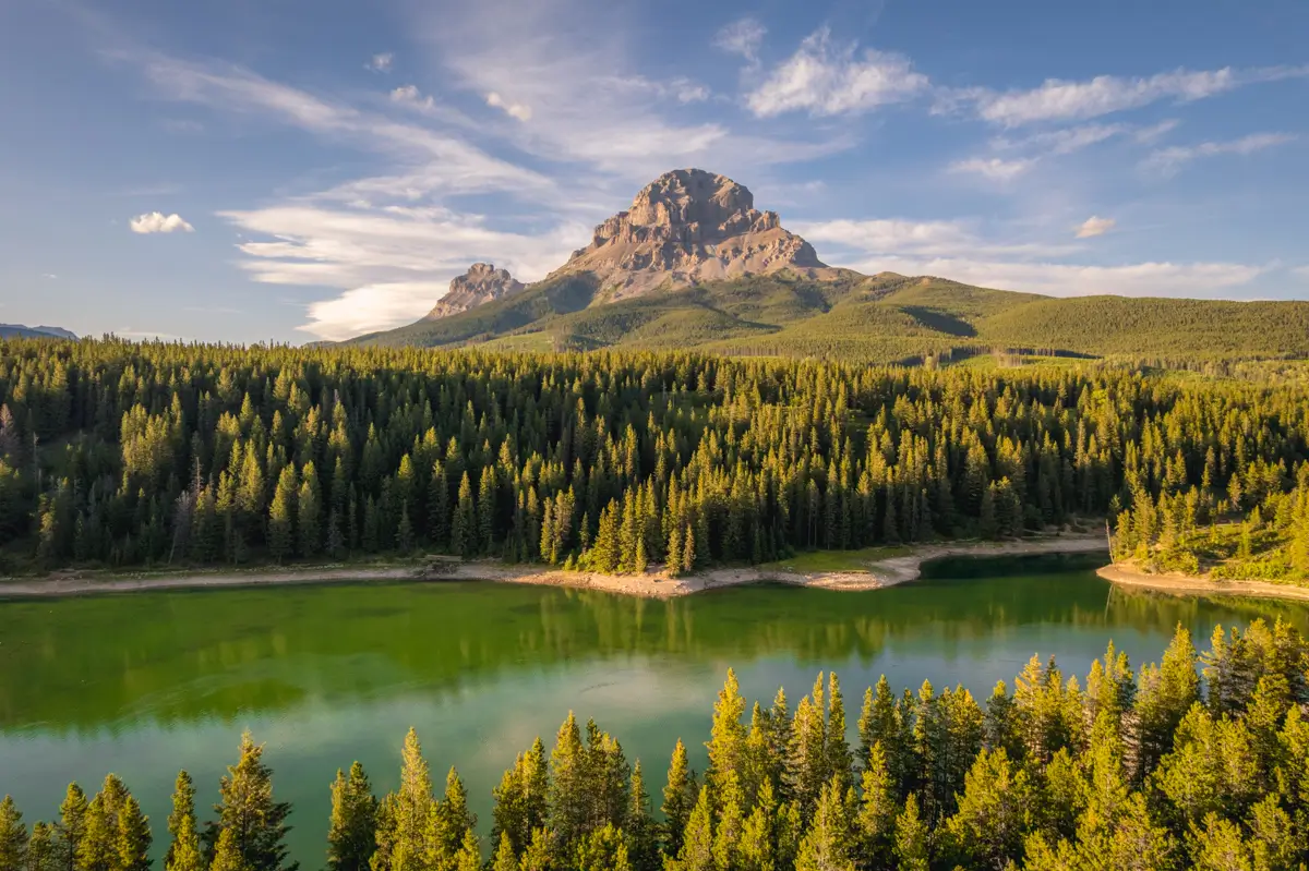 A large mountain rises above a dense green forest with a calm lake in the foreground, under a partly cloudy blue sky near Crowsnest Pass.