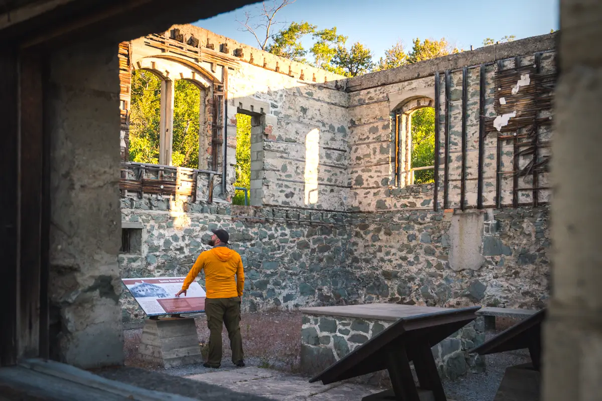 A person in a yellow jacket stands inside the stone ruins of a building in Crowsnest Pass, reading an informational sign. The structure has tall, empty window frames and partial walls with trees visible outside.