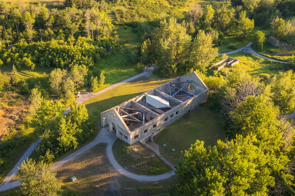 Aerial view of a roofless stone building ruin in Crowsnest Pass, surrounded by green trees and grassy areas, with several walking paths curving around the structure on a sunny day.