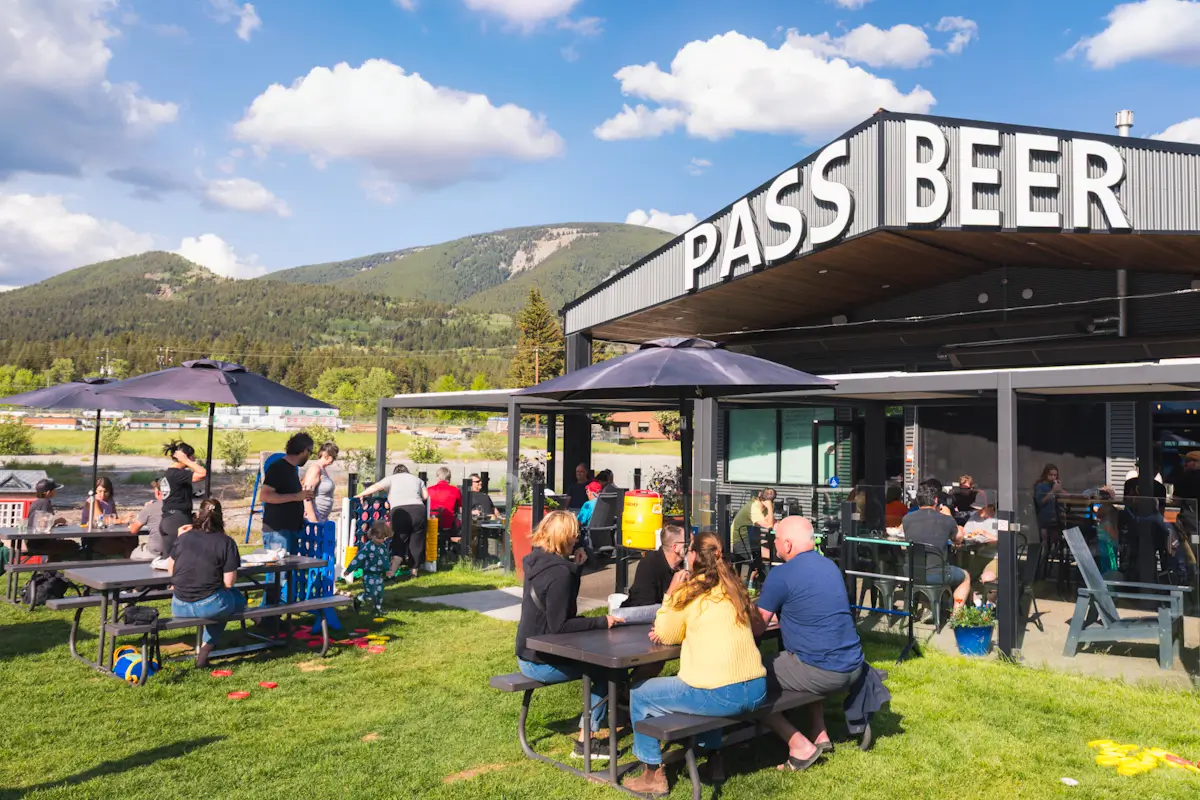People sit at picnic tables on a grassy outdoor patio under umbrellas near a building with a large PASS BEER sign, enjoying views of the Crowsnest Pass mountains and trees in the background beneath a partly cloudy sky.