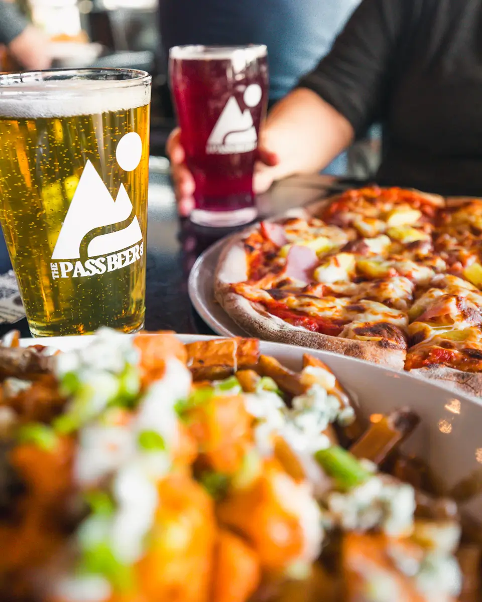 Two people sit at a table in Crowsnest Pass, enjoying pizza, a pint of light beer, a glass of red beverage, and a dish topped with green onions and blue cheese. Both glasses feature The Pass Beer logo.