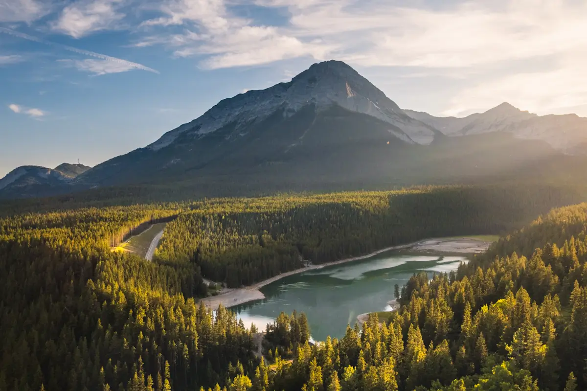 A dense forest surrounds a small lake in the Crowsnest Pass, with a mountain in the background under a partly cloudy sky at sunset or sunrise. Sunlight highlights parts of the trees and mountain slopes.