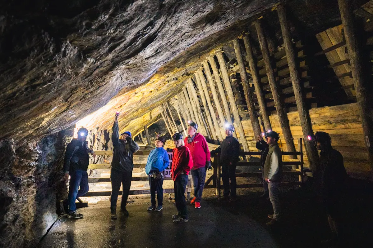 A group of people wearing headlamps stands inside a dimly lit underground Crowsnest Pass mine with wooden supports and rock walls, while one person points upwards.