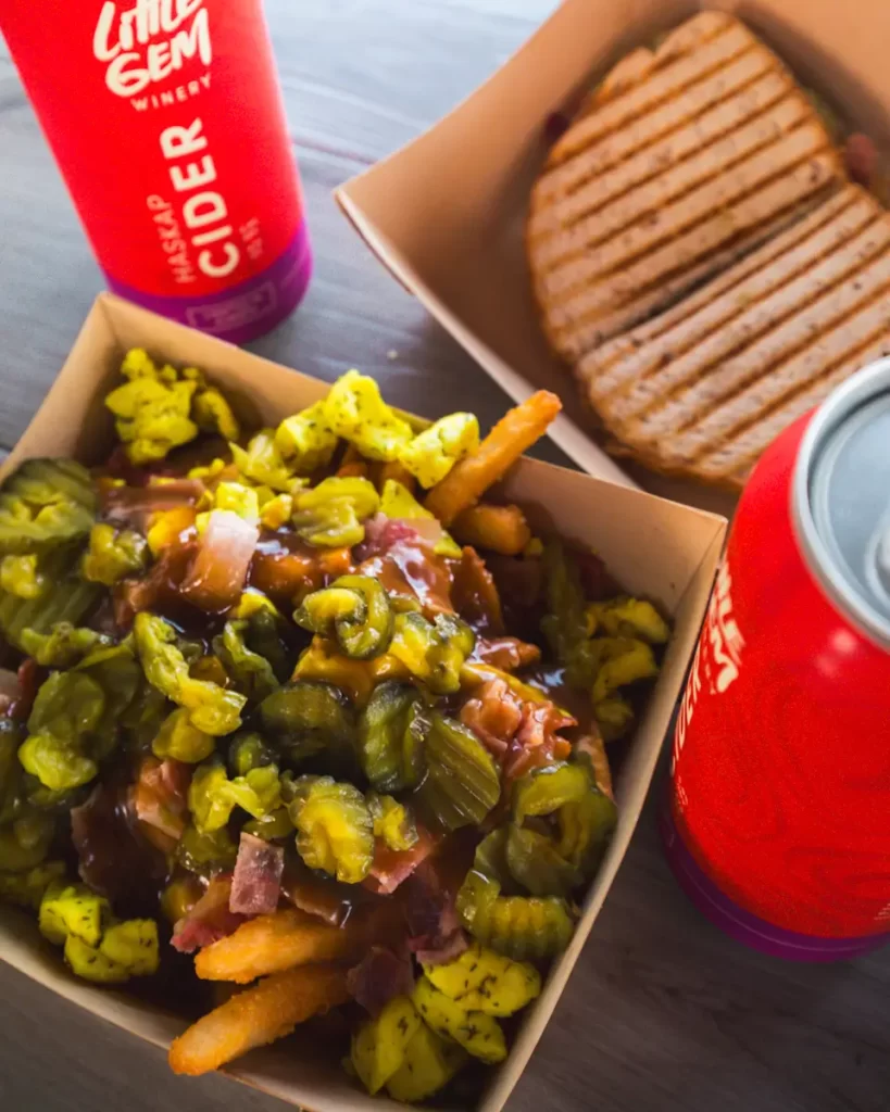 A cardboard container of fries topped with chopped pickles, bits of meat, and brown sauce sits next to a grilled panini sandwich and two cans labeled “Little Gem Winery Cider”—a tasty stop in any Lethbridge guide for local eats.