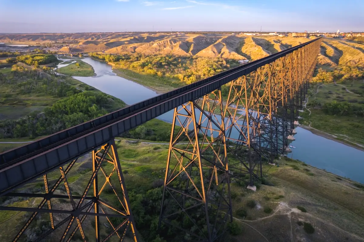 A long, black steel railway bridge crosses over a wide river and green valley, supported by tall metal pillars. Rolling hills and a distant town highlight this iconic spot among top Lethbridge attractions under a bright blue sky.
