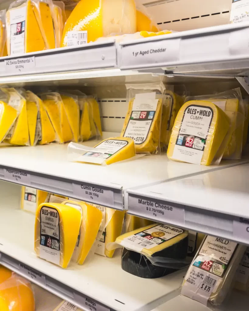 Shelves in a refrigerated grocery store section display various blocks and wedges of cheese in yellow packaging, with labels indicating types such as cumin gouda, marble cheese, and aged cheddar—a tasty stop for any Lethbridge guide.