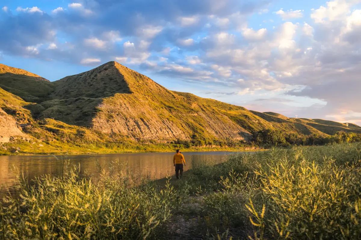 A person in a yellow shirt stands near the edge of a calm river with grassy hills and mountains behind them—one of the scenic Lethbridge attractions featured at sunset under a partly cloudy sky.