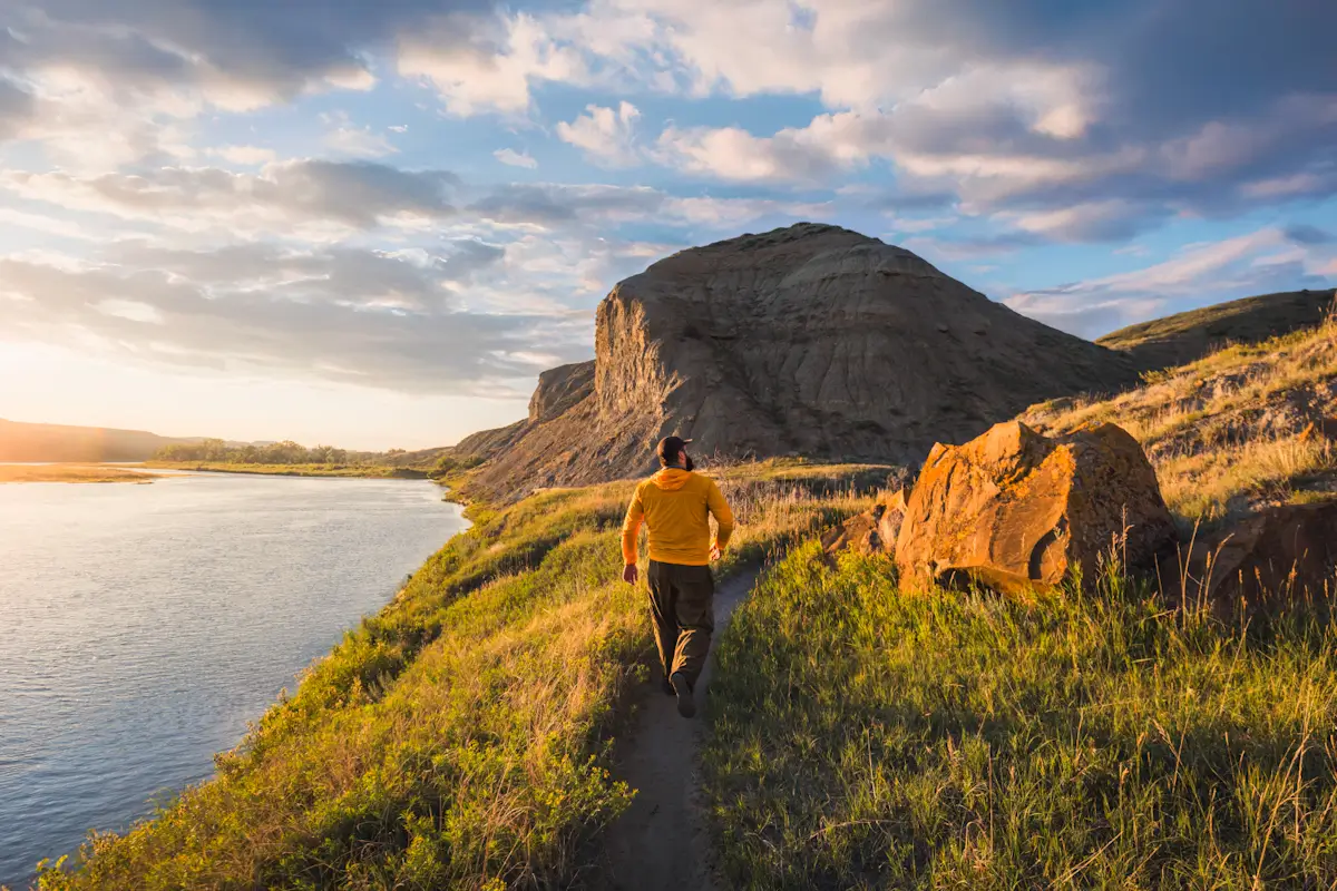 A person wearing a yellow jacket walks along a narrow dirt path beside a river, exploring grassy terrain and large rock formations under a partly cloudy sunset—one of many scenic experiences featured in any Lethbridge guide.