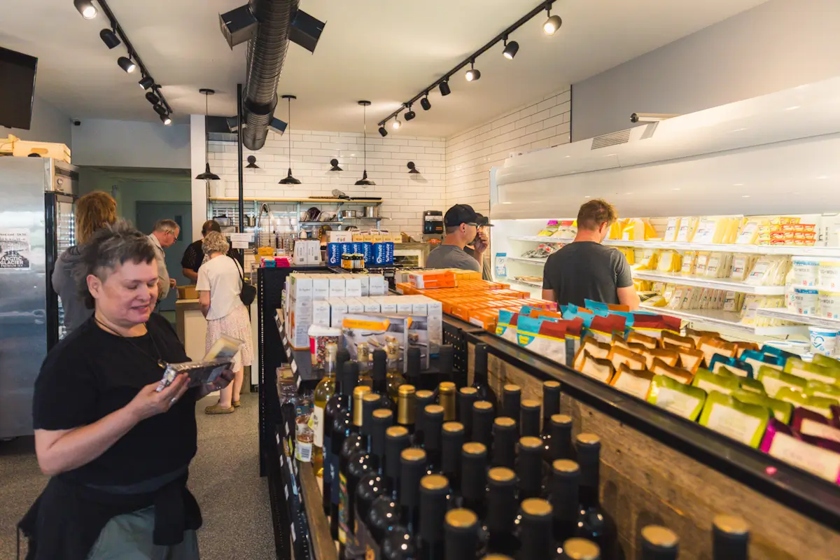 Several people shop in a grocery store, browsing shelves stocked with packaged foods, bottles, and refrigerated products under bright lighting. A woman in the foreground examines an item—an everyday scene among the things to do in Lethbridge.