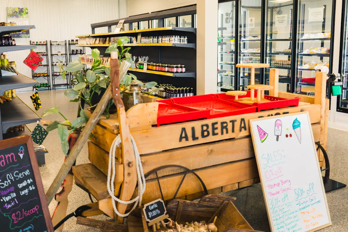 A wooden cart labeled ALBERTA holds produce in a small grocery store—one of many charming Lethbridge attractions. Shelves lined with jars and bottles flank the walls, while a whiteboard sign displays sales and prices near the cart.