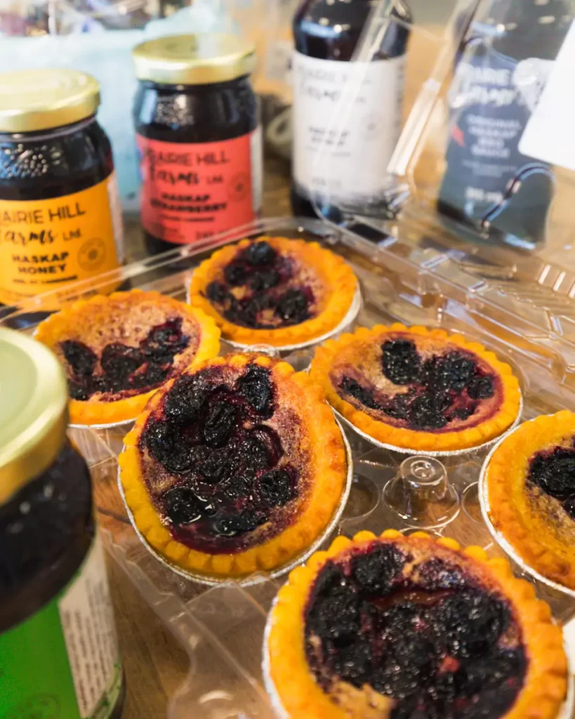 Six small berry tarts in foil cups sit in a plastic container, surrounded by jars of jam and bottles of syrup on a wooden surface—a sweet highlight among Lethbridge attractions for any foodie exploring the city.