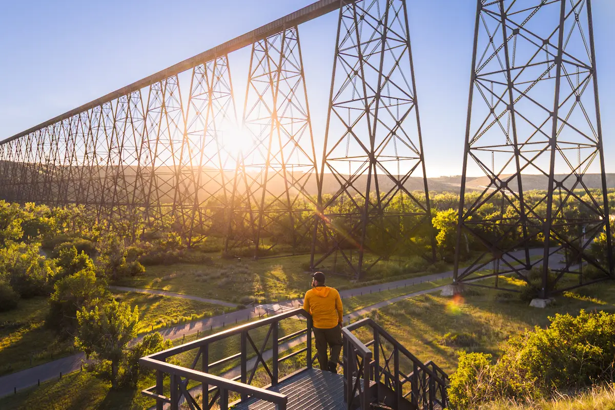 A person in a yellow jacket stands on a wooden platform, admiring a tall steel railway bridge—one of the top Lethbridge attractions—stretching across a green landscape as the sun shines brightly in the background.