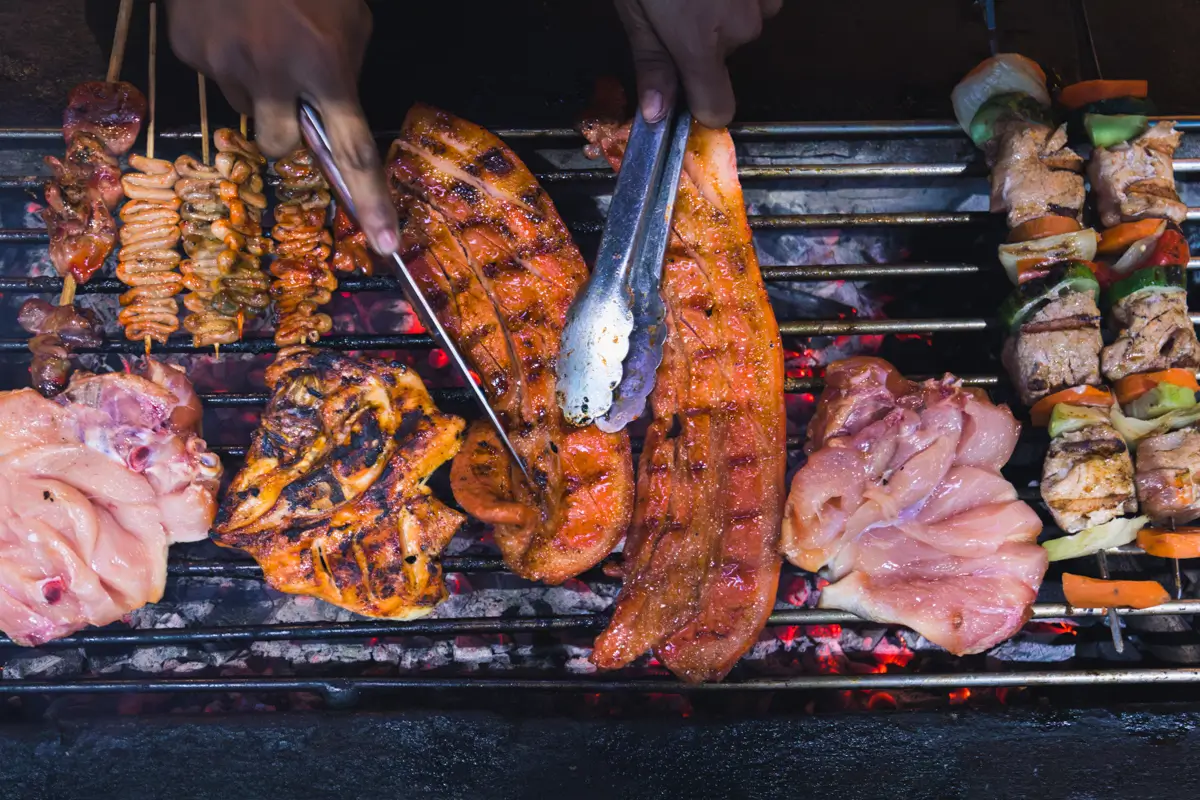 A close-up view of various meats, including skewers, chicken pieces, and ribs, being grilled over hot coals in Moalboal, with hands using tongs and a fork to turn the food.