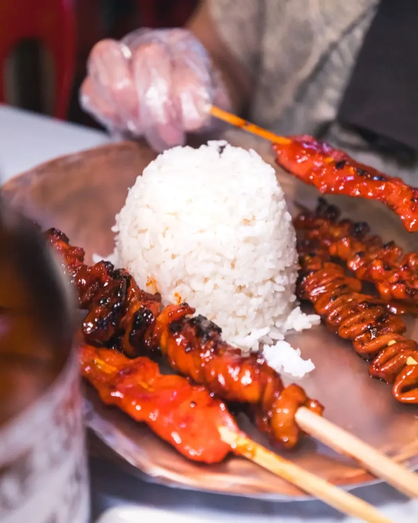 A plate with a mound of white rice in the center, surrounded by several skewers of grilled meat and intestines—a classic Moalboal street food. A person wearing a plastic glove is holding one of the skewers, ready to take a bite.