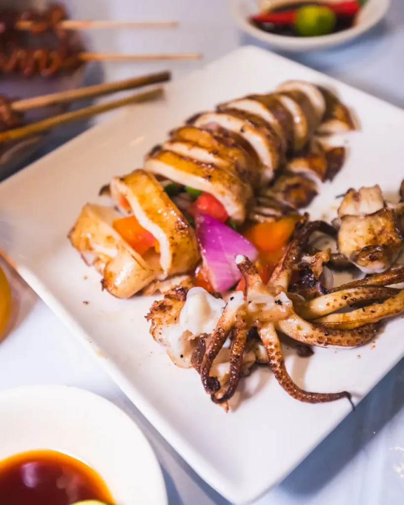 Grilled squid sliced and arranged on a white square plate, garnished with vegetables—a delightful taste of Moalboal. Dipping sauces and other skewered food items are visible in the background.