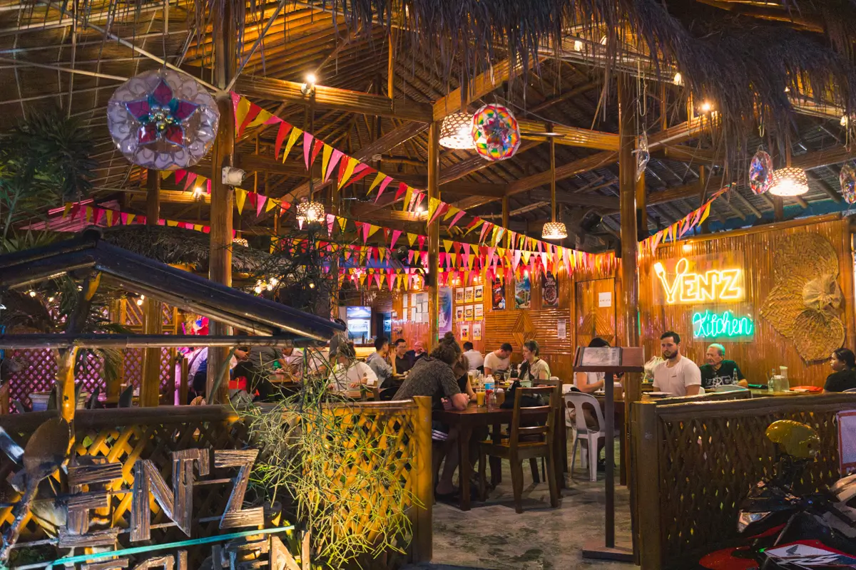 A lively restaurant in Moalboal with wooden decor, colorful flags, and neon signs. Several people are seated at tables, eating and talking. Warm lighting creates a cozy atmosphere, while plants and hanging decorations add to the festive setting.