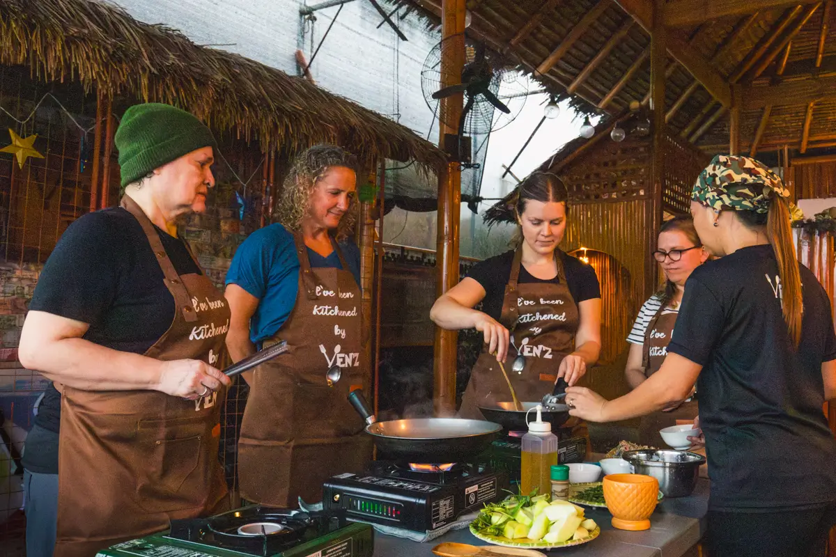 Five people wearing aprons stand around a cooking station under a thatched roof in Moalboal, preparing food together. Several ingredients and cooking utensils are arranged on the table in front of them.