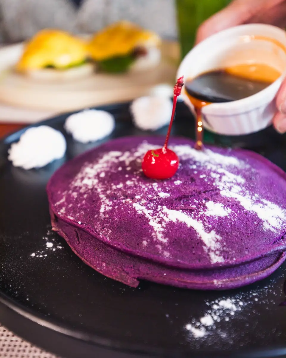 A hand pours syrup onto a stack of purple pancakes topped with a cherry and powdered sugar on a black plate, with dollops of cream beside it—a delightful Moalboal-inspired breakfast scene.