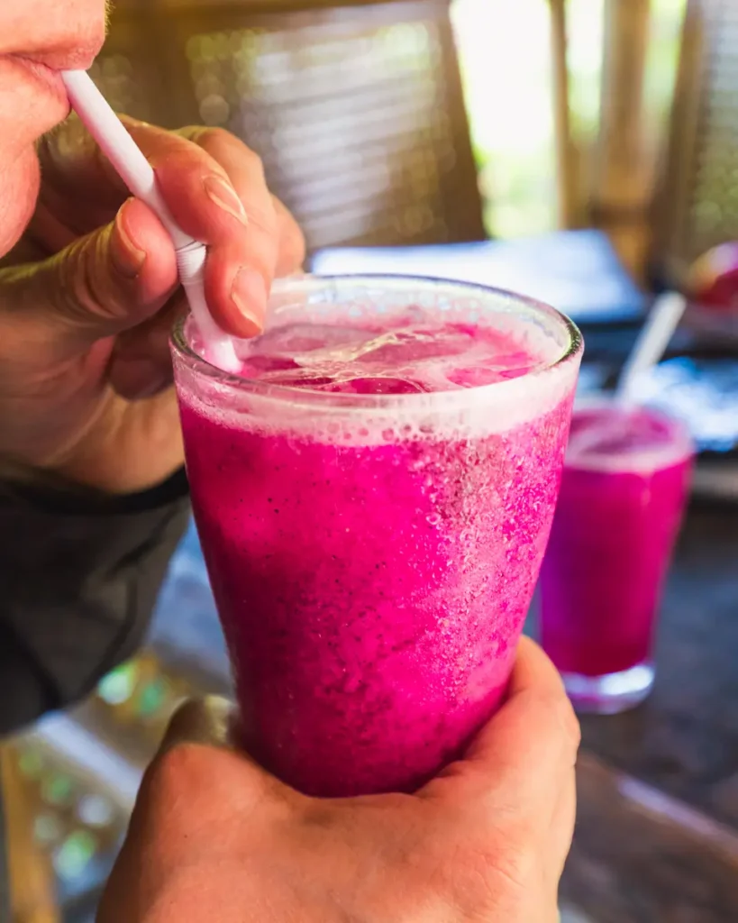 A person enjoys a glass of bright pink dragon fruit juice with ice through a white straw in Moalboal, with another similar glass in the background on a wooden table.