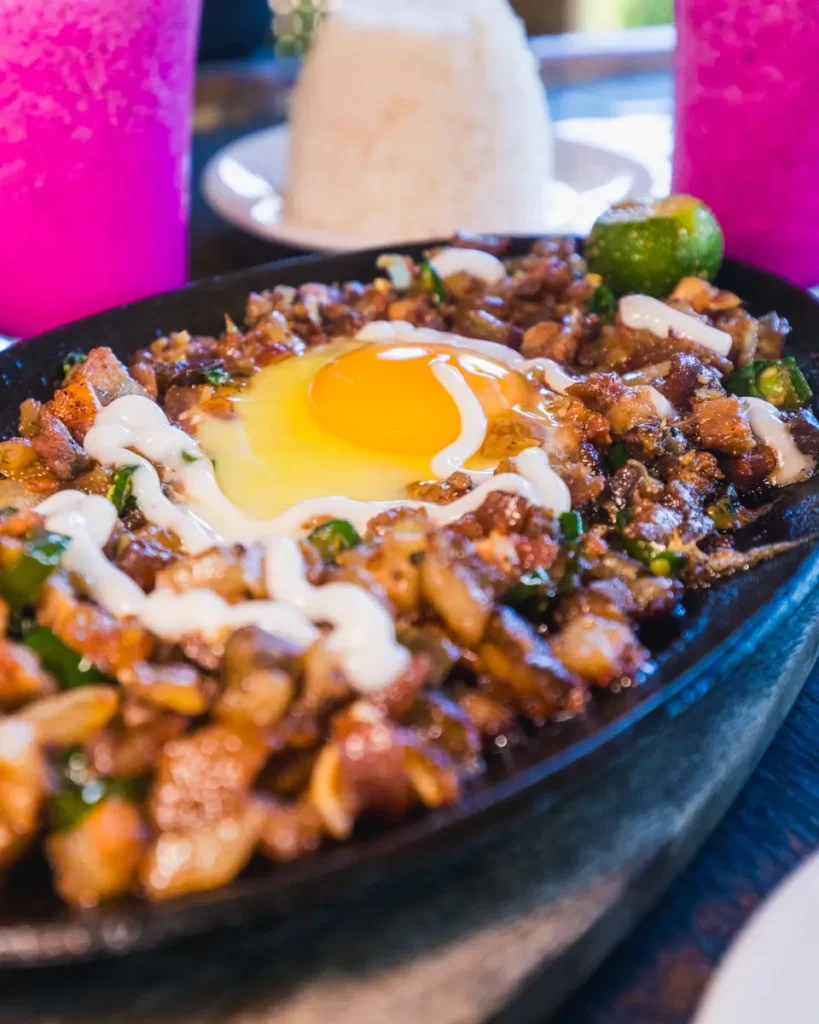 A sizzling plate of chopped meat topped with a raw egg and drizzled with white sauce, served Moalboal-style, with a mound of rice and two pink drinks in the background.