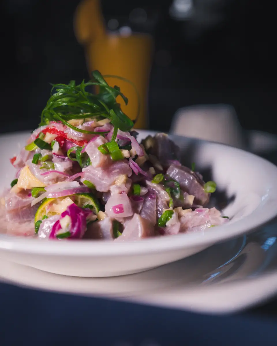 A close-up of a white bowl filled with a Moalboal-inspired salad featuring pieces of fish, red onions, green onions, and herbs, garnished with sliced green onions. The background is dark and out of focus.