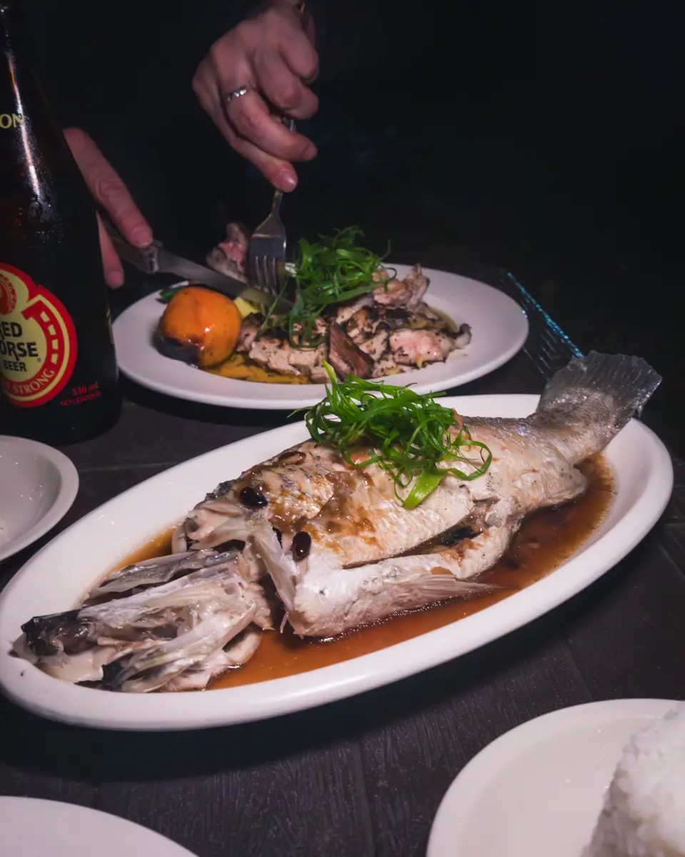 A cooked whole fish, served Moalboal-style on an oval white plate with sauce and green garnish, sits next to another dish being sliced, a beer bottle, and a bowl of rice. Two hands are seen using a fork and knife.
