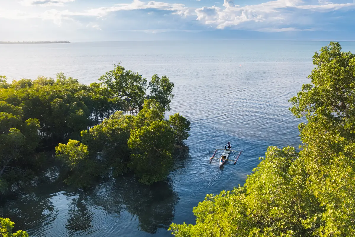 Two people paddle a canoe through calm blue water in Moalboal, surrounded by green trees, sunlight reflecting on the lake, and clouds drifting in the sky.