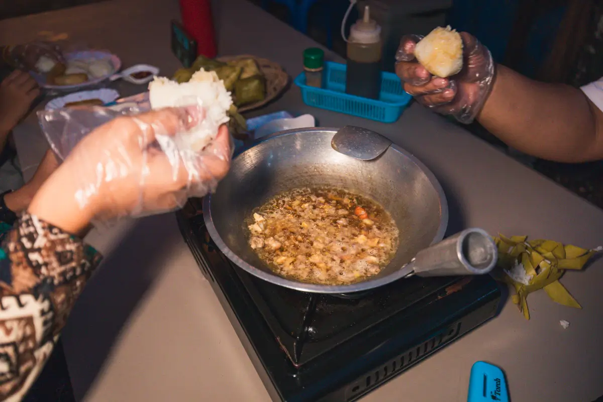 Two people wearing plastic gloves are preparing food over a gas stove in Moalboal. One holds a pot with oil and chopped ingredients, while the other shapes rice. Cooking utensils and condiments are on the table.