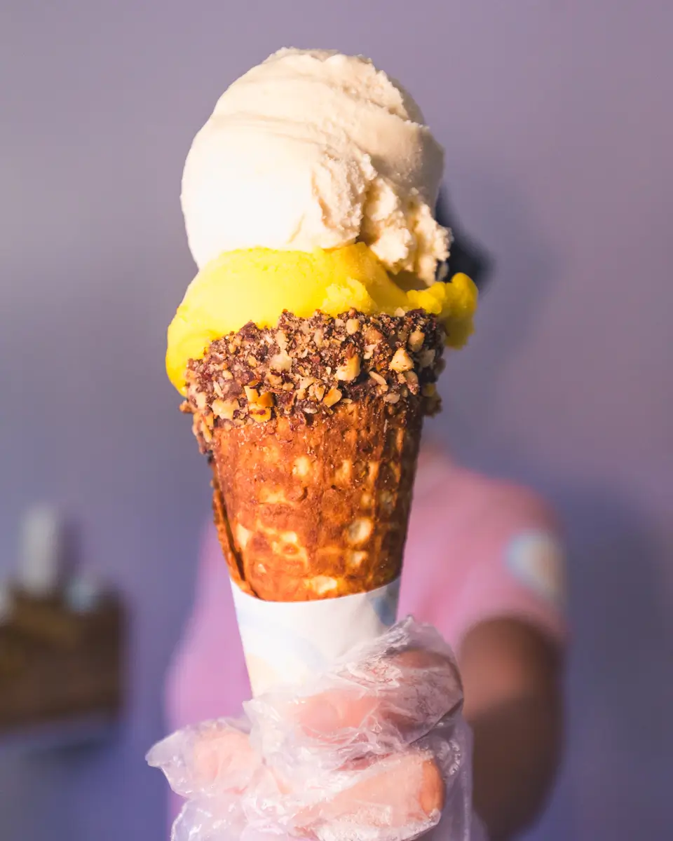 A person wearing a clear glove holds out an ice cream cone with two scoops, one yellow and one white, in a waffle cone dipped in chocolate and nuts. The purple-hued background hints at a vibrant day in Moalboal.