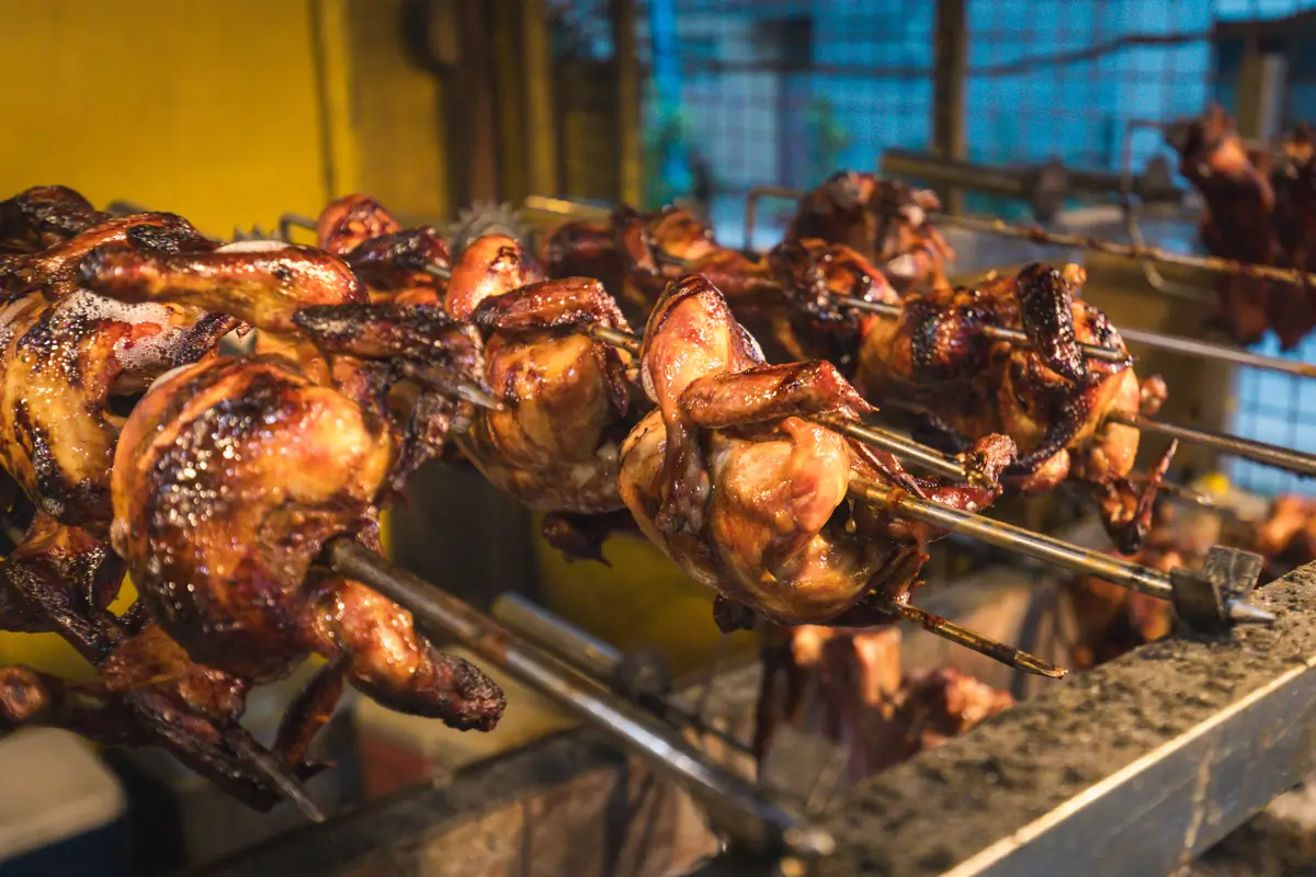 Whole chickens are skewered on metal rods and roasting over an open flame in a rotisserie grill setup in Moalboal. The chickens appear golden brown and cooked, with a yellow wall and metal grating in the background.
