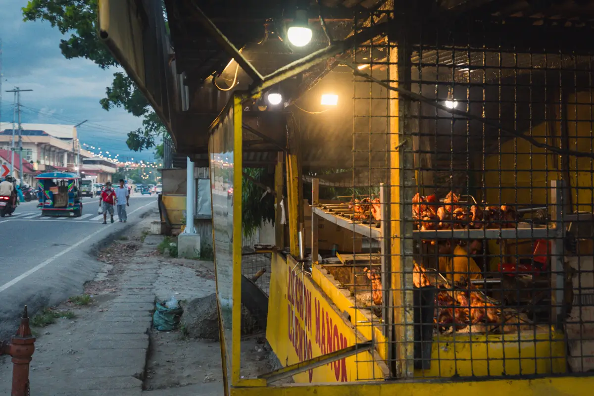 A sidewalk scene in Moalboal shows a roadside food stall with roasted chickens displayed behind a metal grate under warm lights. A yellow counter is visible, and people and vehicles are seen in the background alongside the street.