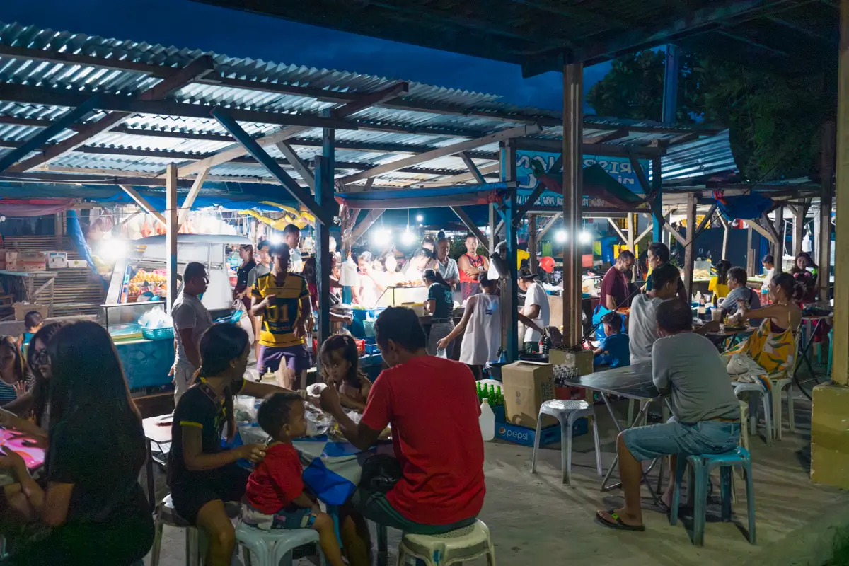 People gather at an outdoor market in Moalboal at night, sitting at tables and eating food under covered stalls with bright lights. The area is busy, with vendors and customers interacting in a lively atmosphere.