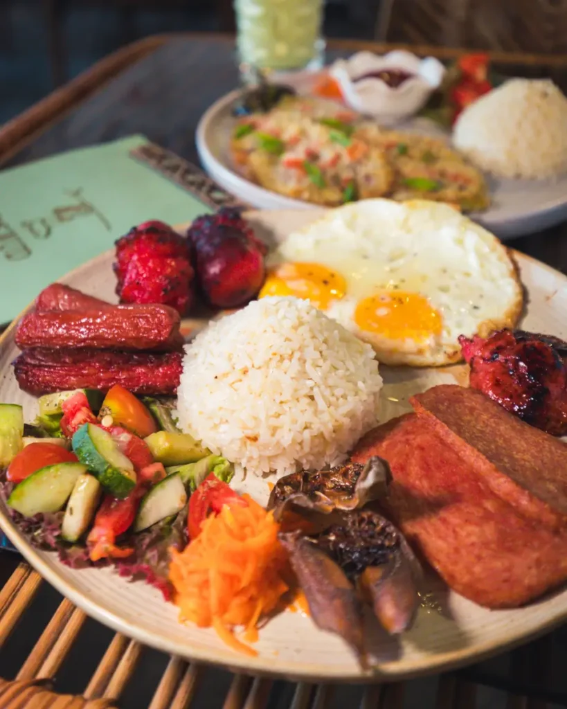 A plate of Filipino breakfast food in Moalboal featuring white rice, fried eggs, grilled meat, longganisa sausages, fried spam, dried fish, mixed vegetables, and pickled shredded carrots, with another meal in the background.