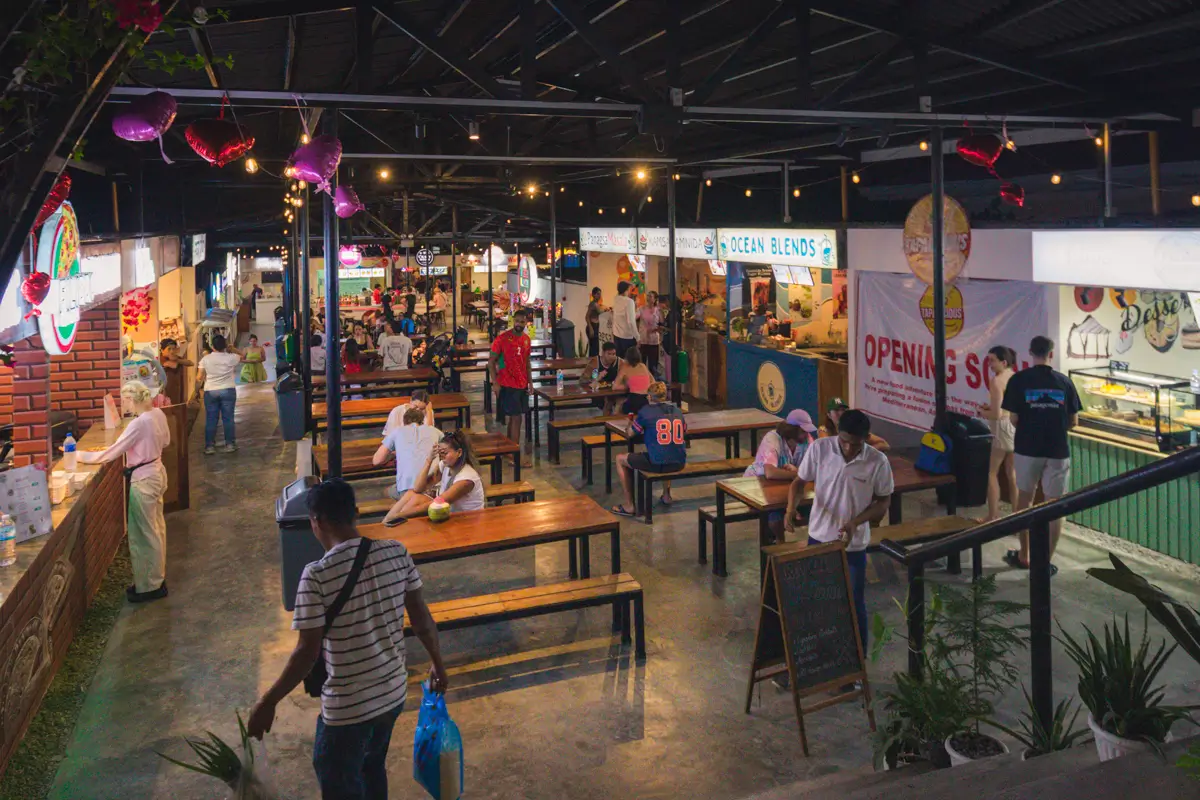 Indoor food court in Moalboal with several wooden tables and benches, people sitting, standing, and walking around, plus various food stalls along the sides with bright signs, under an exposed ceiling and hanging lights.