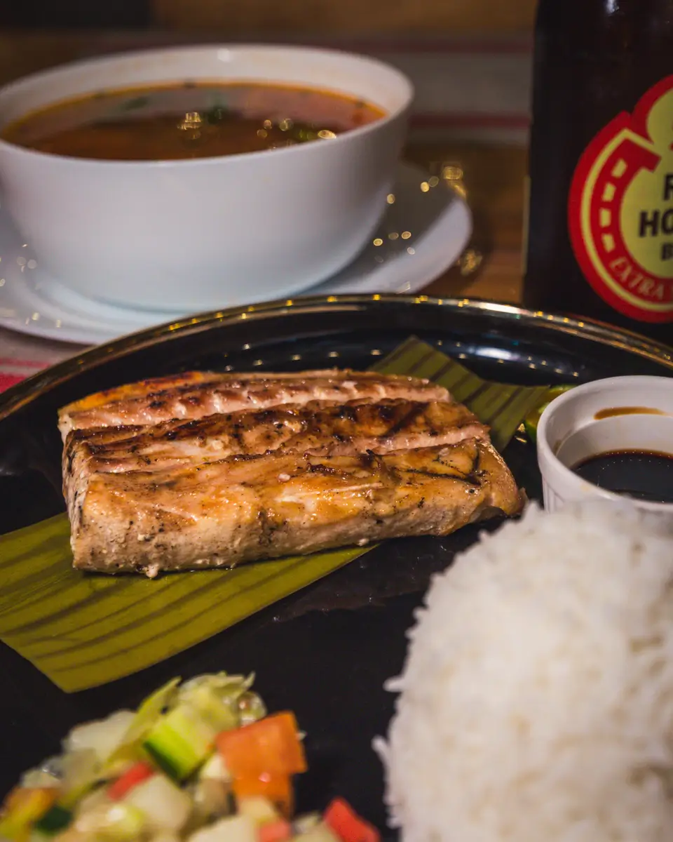 A plate with a grilled fish fillet on a banana leaf, white rice, sauce, and vegetable salad captures the flavors of Moalboal. In the background, a bowl of soup and a bottle of beer complete this inviting local meal.