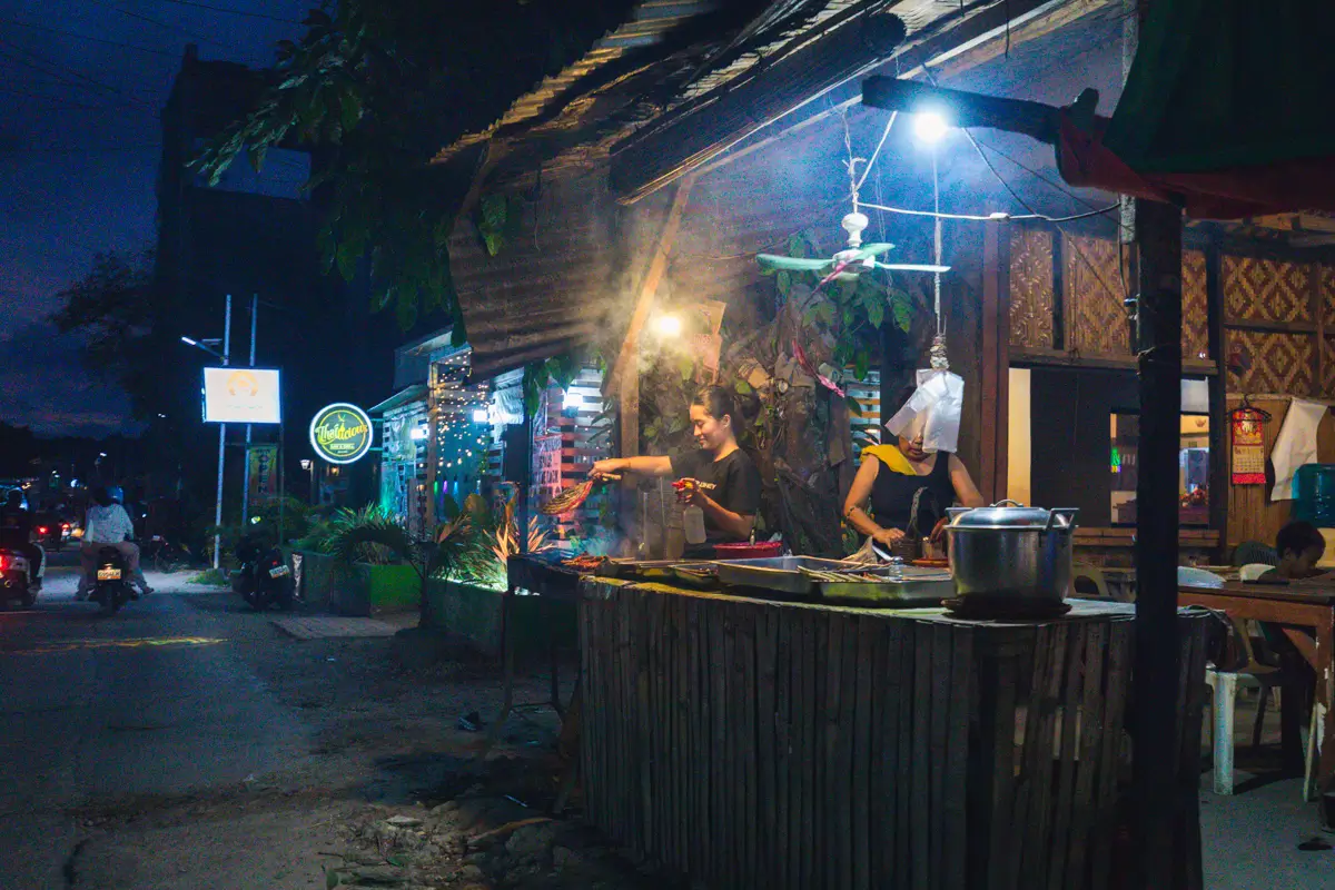 A woman cooks at a Moalboal outdoor food stall at night, illuminated by hanging lights, with a pot in the foreground and another person preparing food in the dimly lit street and storefronts.