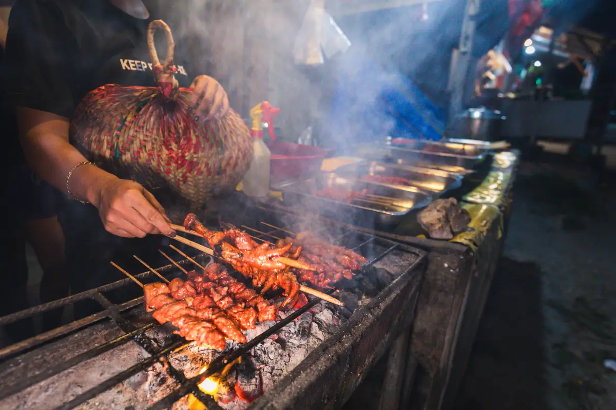 At an outdoor food stall in Moalboal, a person grills skewers of meat over charcoal, smoke rising as trays of food fill the background.