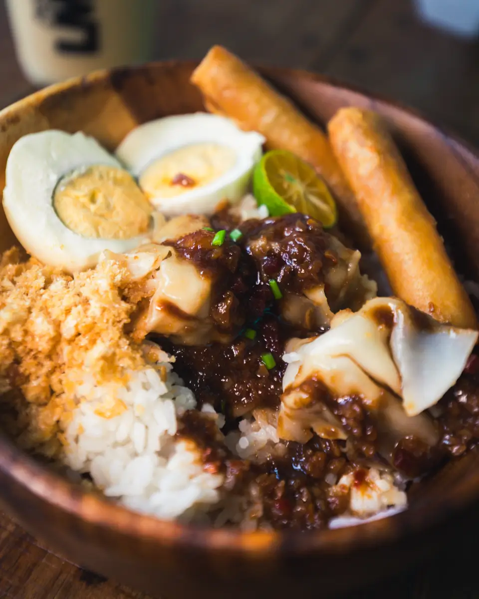 A wooden bowl filled with white rice, hard-boiled egg halves, lumpiang shanghai, siomai topped with brown sauce, garlic bits, and a slice of calamansi sits on a wooden table—bringing the taste of Moalboal to your meal.