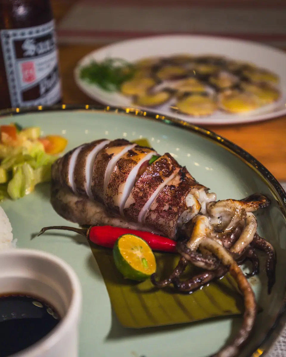 A plate with a grilled whole squid, sliced and served with a small salad, a red chili pepper, a lime wedge, and dipping sauce—capturing the fresh coastal flavors of Moalboal. In the background sits another dish and a bottle.